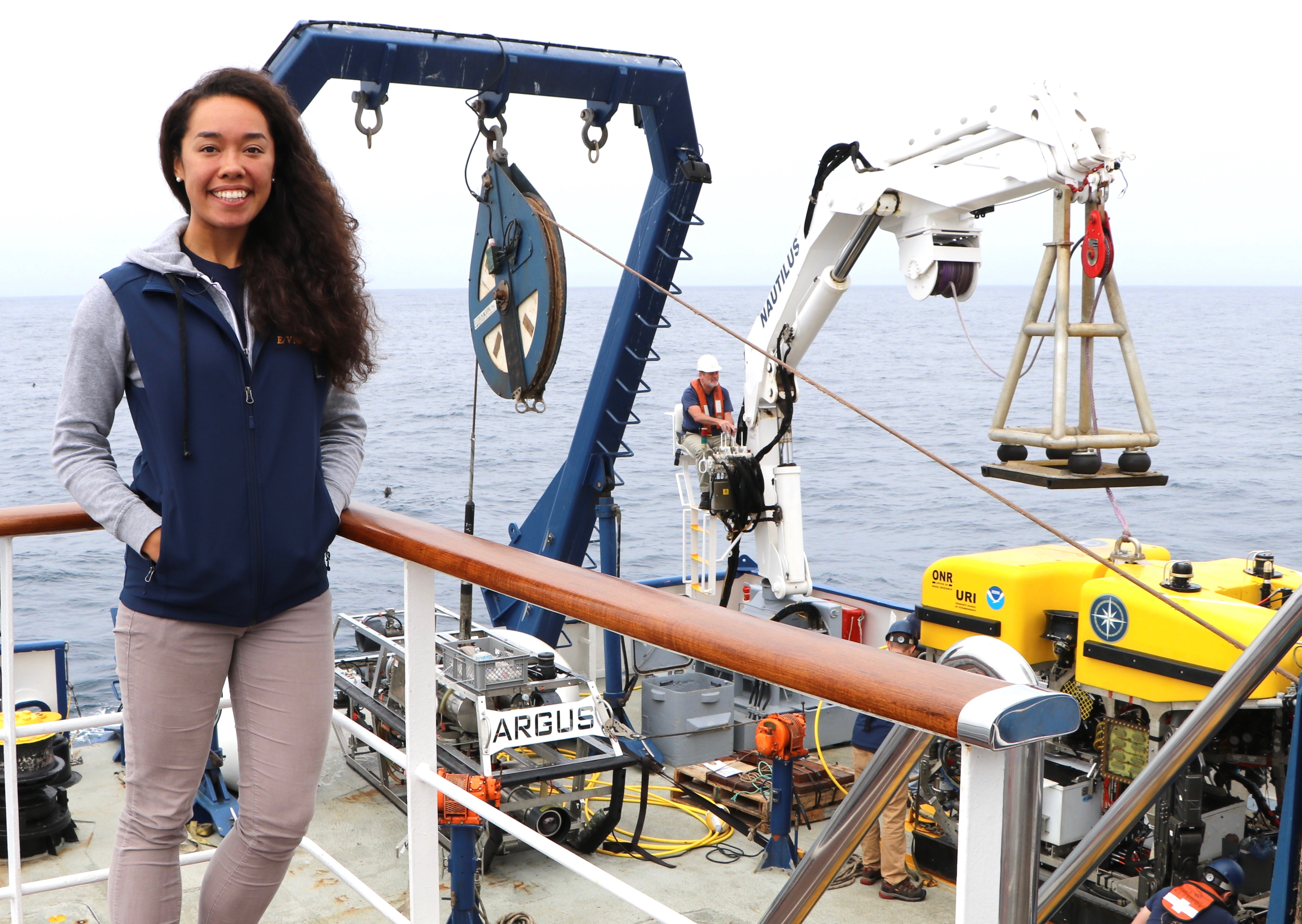 Carina standing on the second deck level of E/V Nautilus with crane and ROV Hercules + Argus in background.